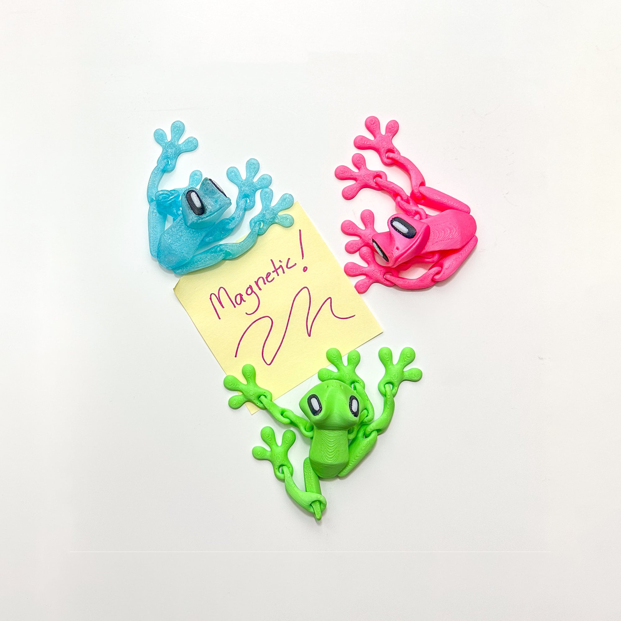 Blue, pink and green 3D Printed Magnetic Frog Locker Buddies surrounding a handwritten “Magnetic!” note on a white magnetic surface.